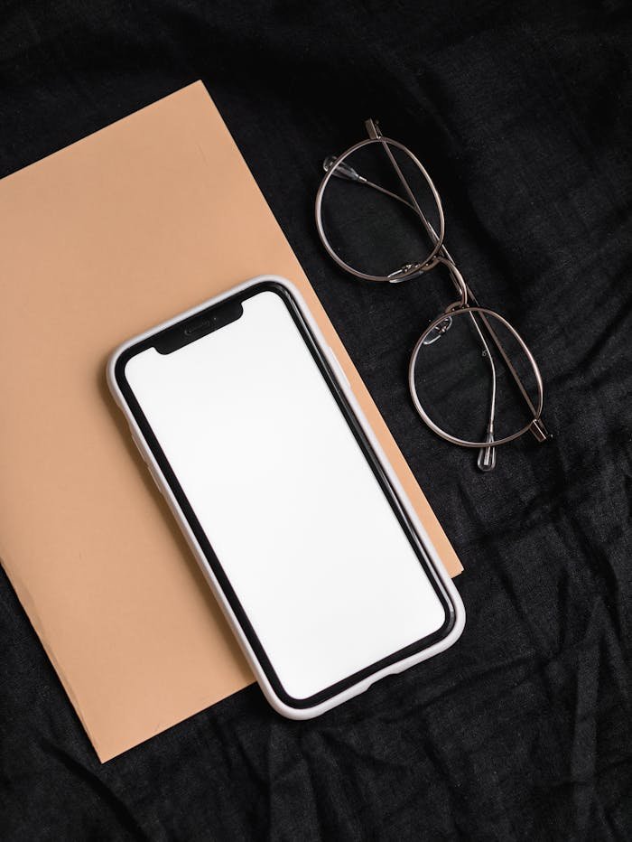 A stylish flatlay featuring a smartphone, eyeglasses, and a notebook on a black surface.